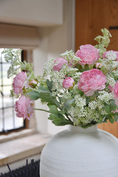 White sphere vase placed on a kitchen countertop with an arrangement of pale pink ranunculuses and white mini bud stems. Displayed in a country style kitchen for home decor.