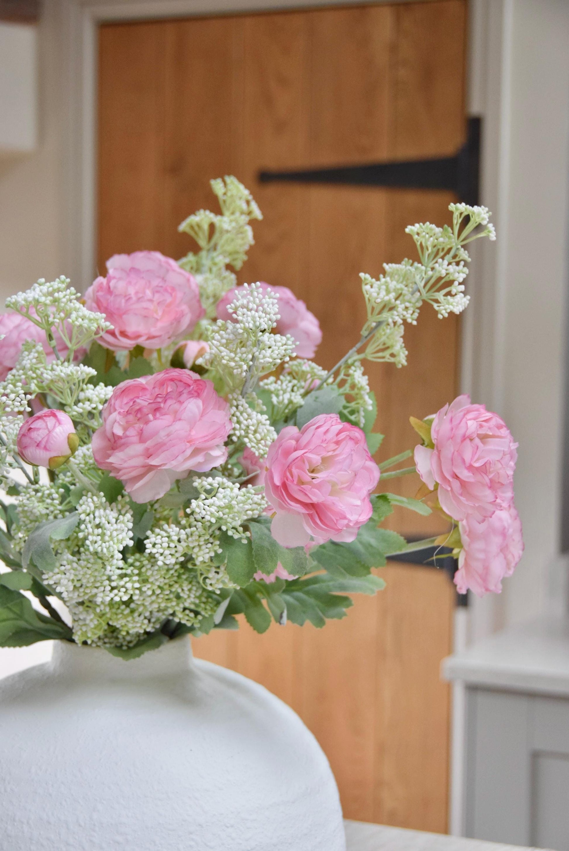 White sphere vase placed on a kitchen countertop with an arrangement of pale pink ranunculuses and white mini bud stems. Displayed in a country style kitchen for home decor.