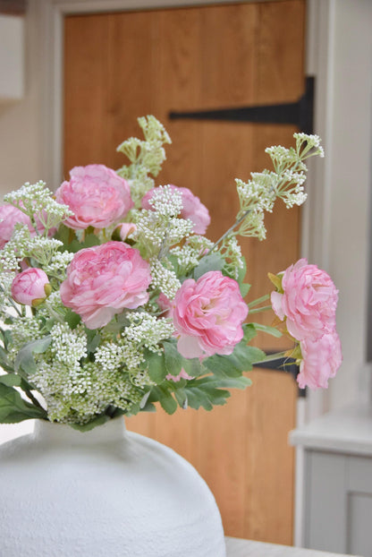 White sphere vase placed on a kitchen countertop with an arrangement of pale pink ranunculuses and white mini bud stems. Displayed in a country style kitchen for home decor.