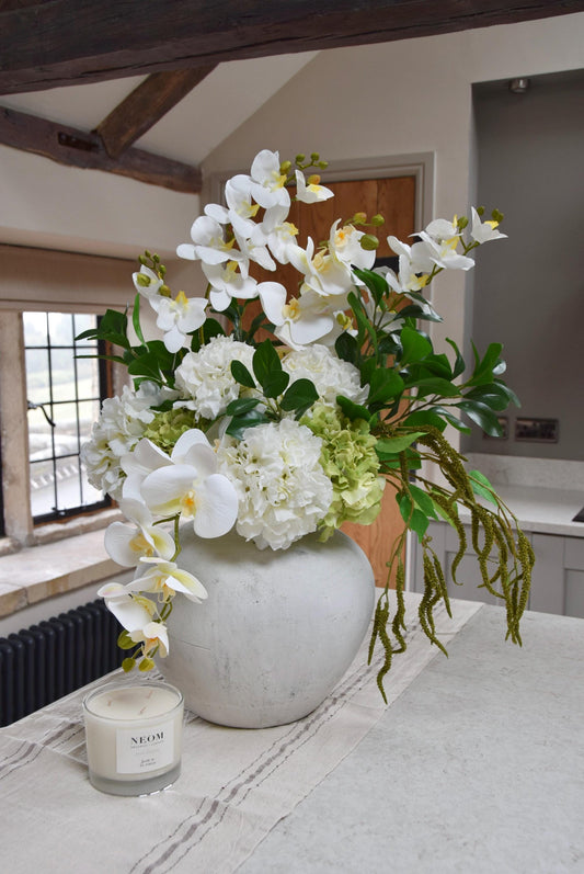 Stone antique vase placed on a kitchen countertop with an arrangement of white orchids, white hydrangeas, green hydrangeas and green foliage. Displayed in a country style kitchen for home decor.