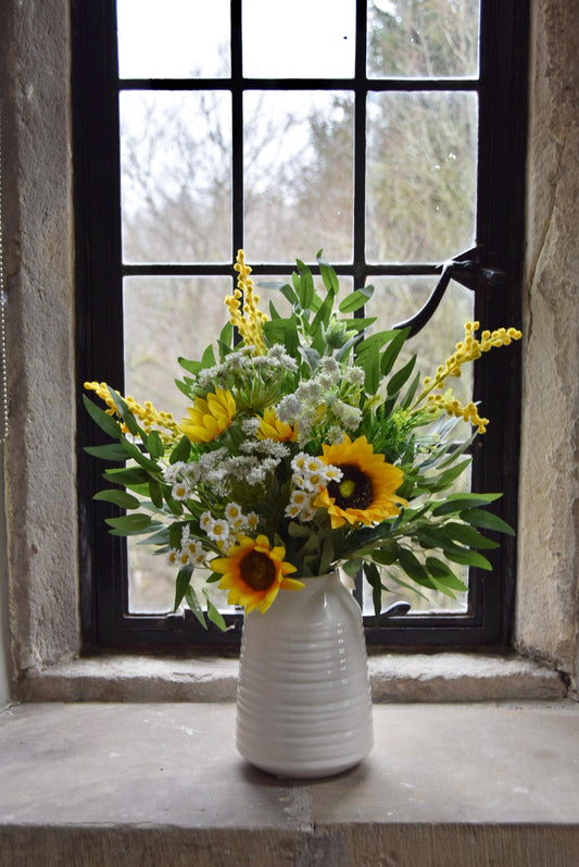 Cream country style vase placed on a stone windowsill with an arrangement of yellow sunflowers and green foliage. Displayed in a country style window for home decor.