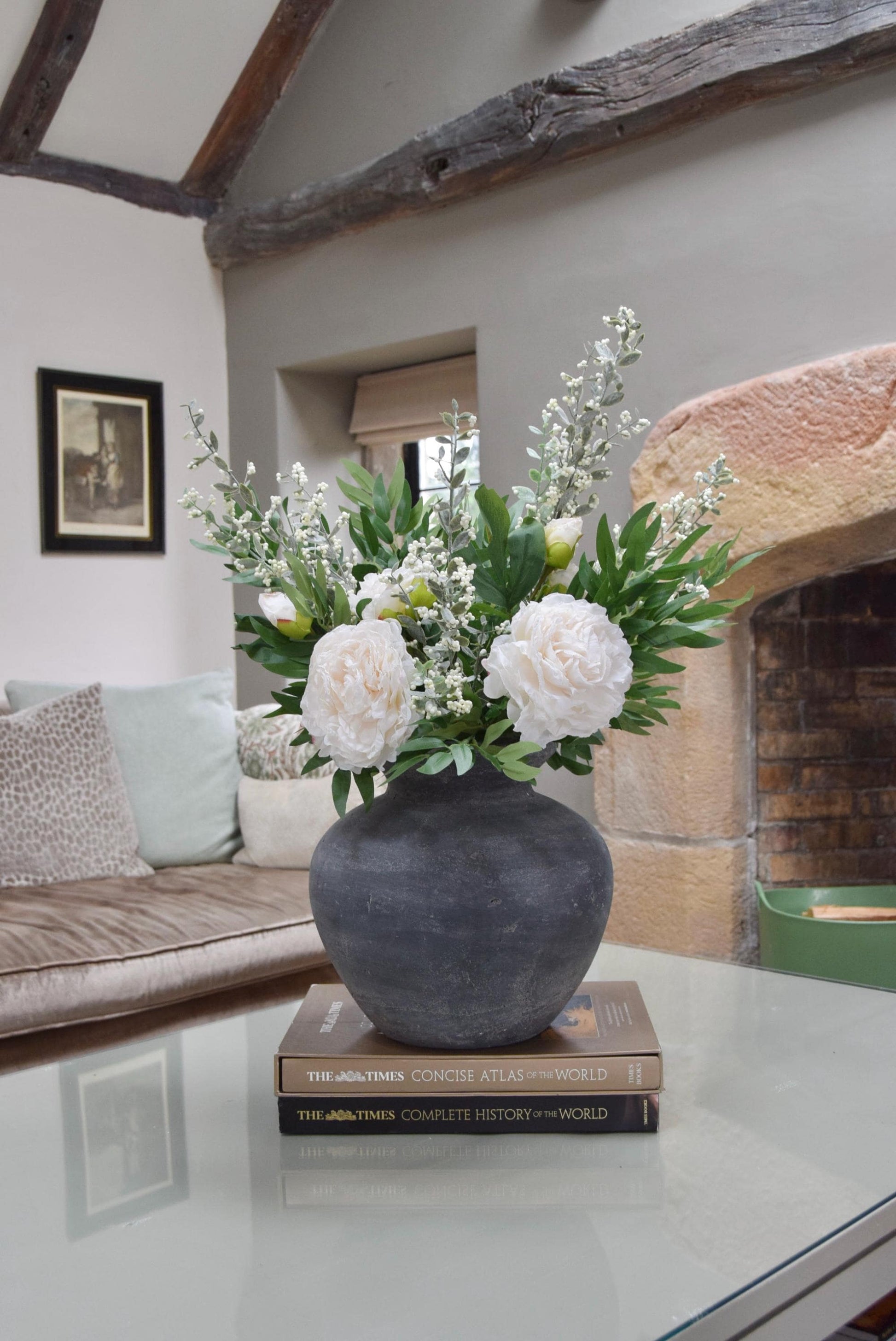 Grey antique vase placed on a coffee table with artificial ruscus foliage and faux cream peony flowers. Arranged in a country style living room for home decor.