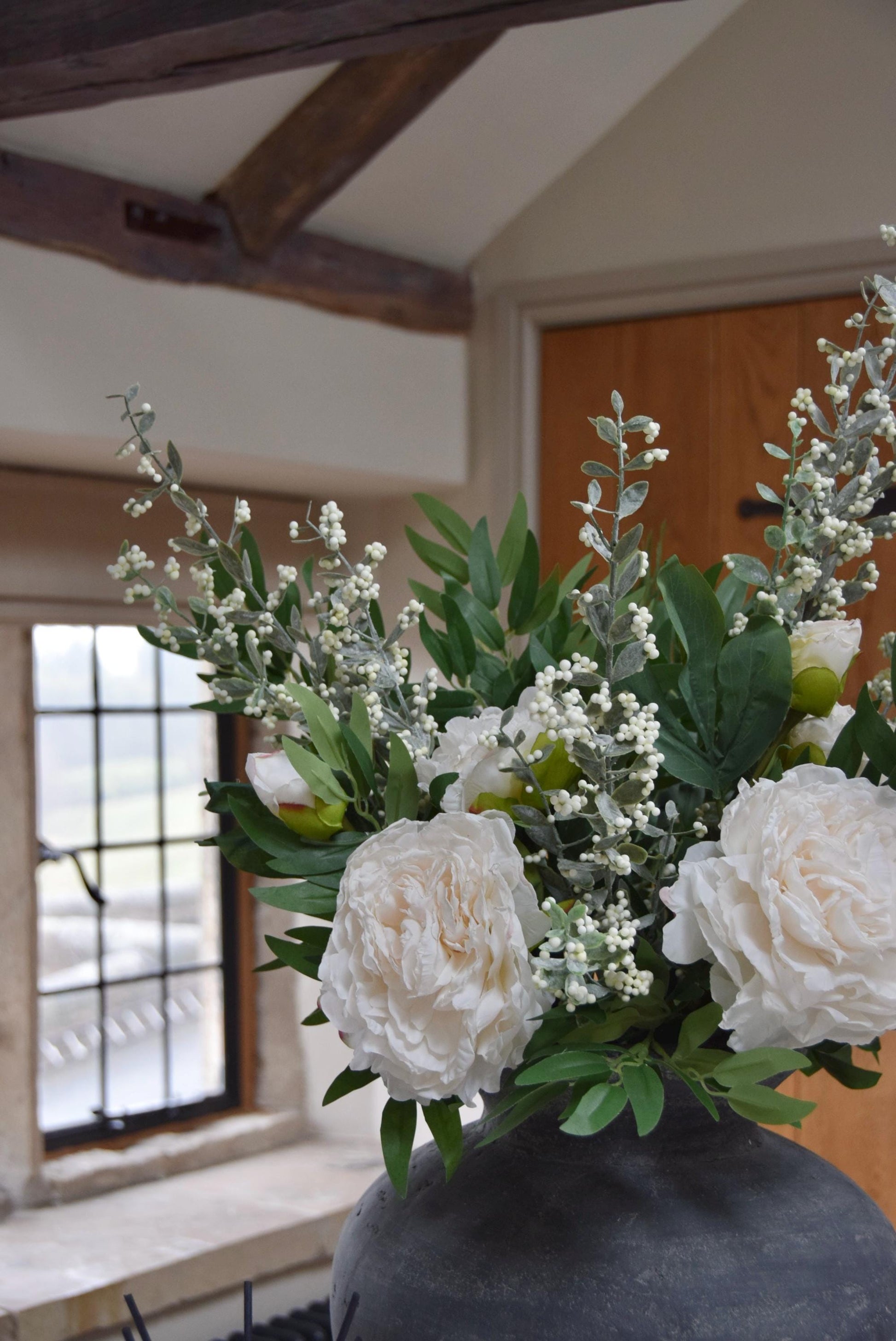 Grey antique vase placed on a kitchen countertop with artificial ruscus foliage and faux cream peony flowers. Arranged in a country style kitchen for home decor.