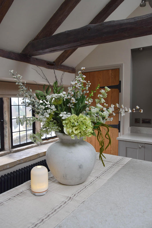 Stone antique vase placed on a kitchen countertop with an arrangement of green and white mixed flowers with lots of foliage. Displayed in a country style kitchen for home decor.