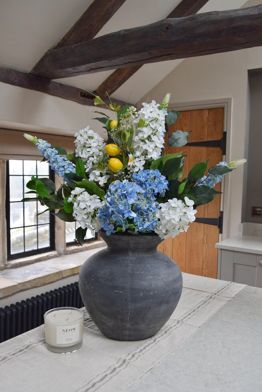 Grey antique vase placed on a kitchen countertop with arrangement of blue and white hydrangeas with lemons and green foliage. Displayed in a country style kitchen for home decor.