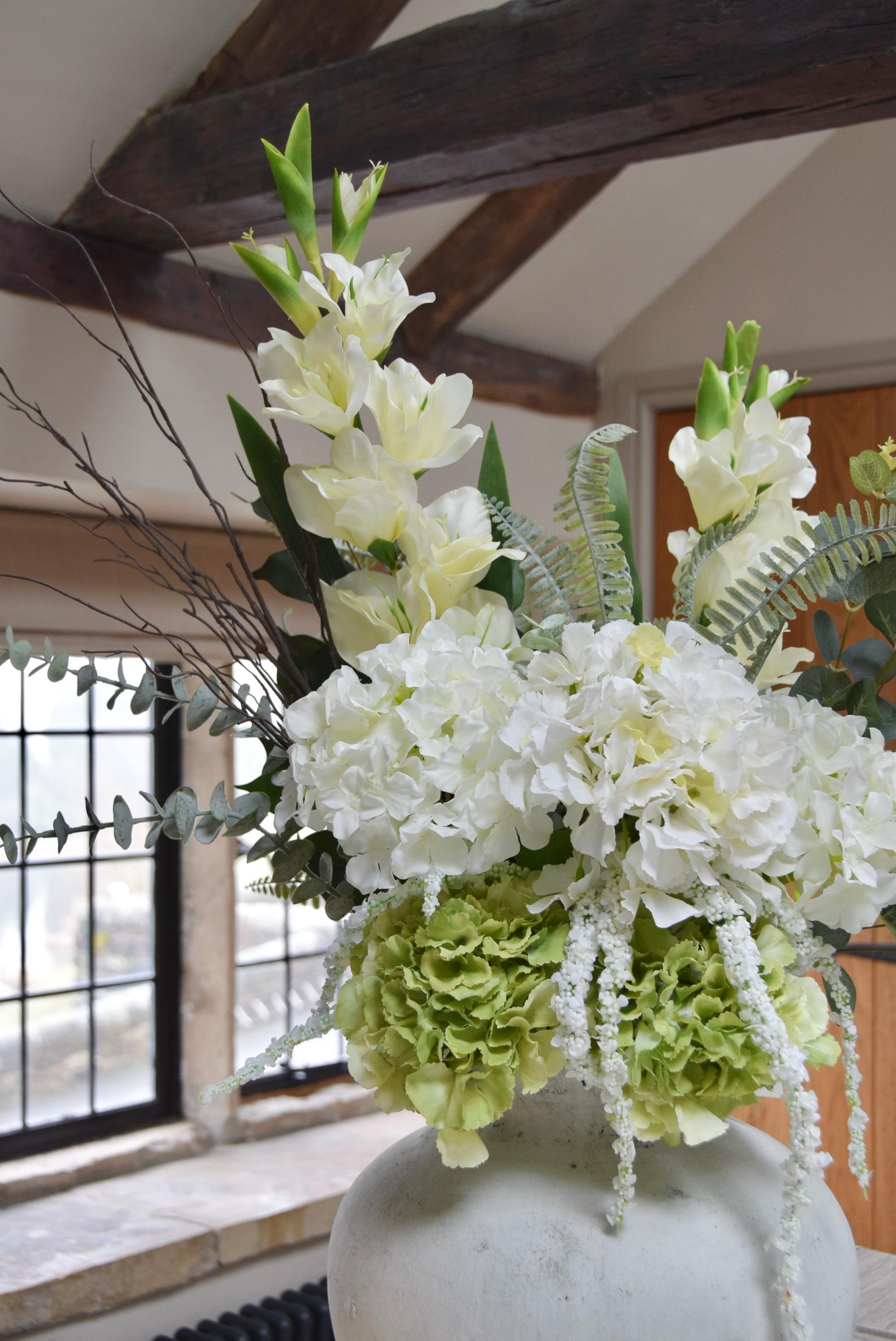 Stone antique vase placed on a kitchen countertop with an arrangement of white hydrangeas, green hydrangeas, cream gladioli and foliage. Displayed in a country style kitchen for home decor.