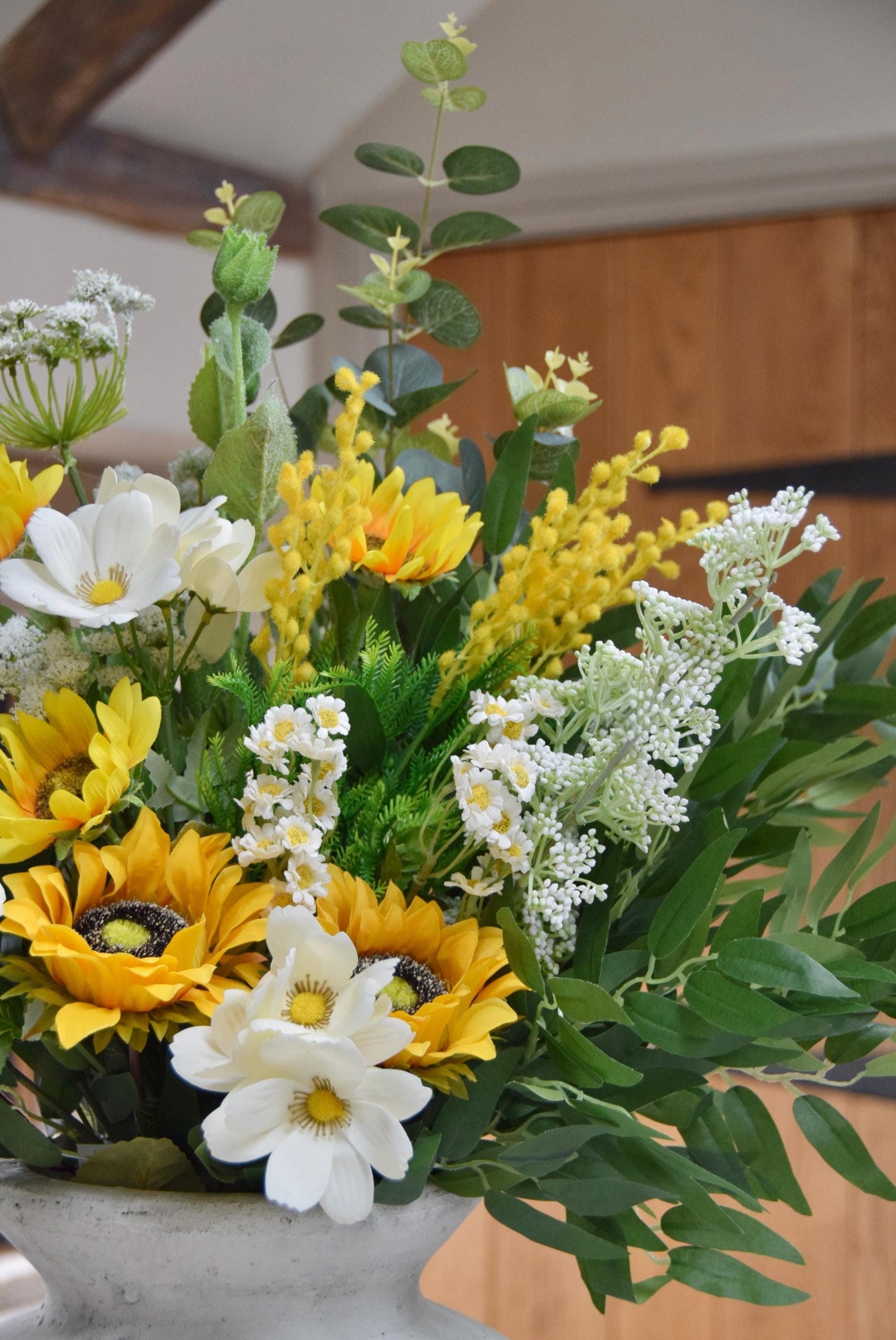 Stone antique vase placed on a kitchen countertop with an arrangement of yellow sunflowers, white wildflowers and green foliage. Displayed in a country style kitchen for home decor.