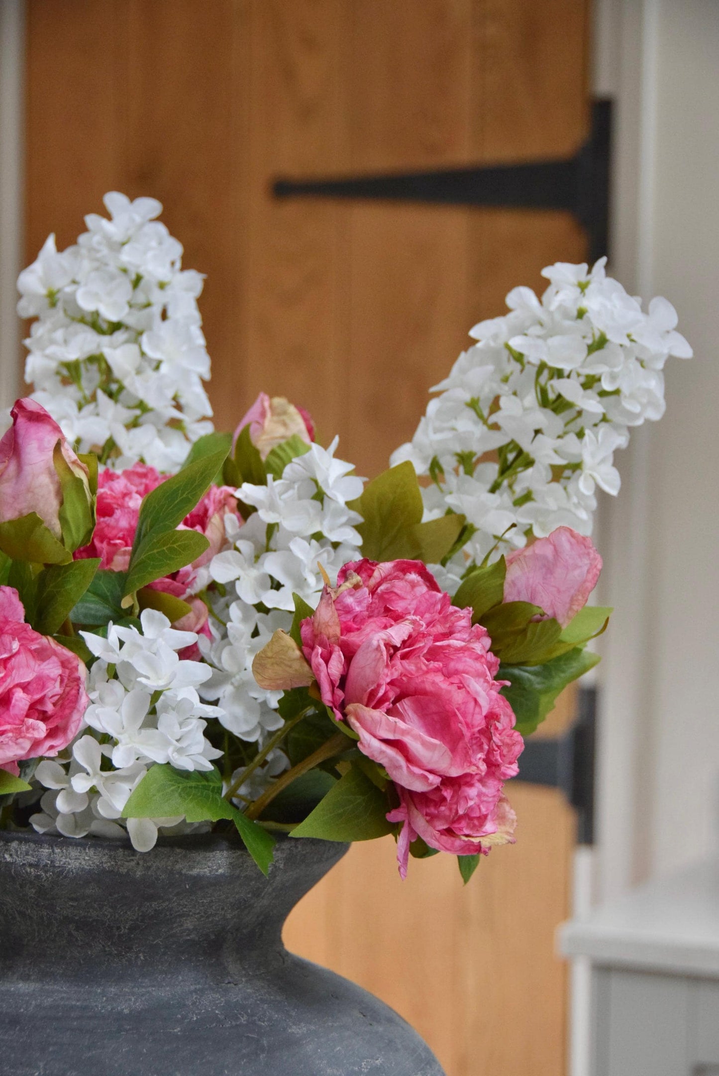 Grey antique vase placed on a kitchen countertop with an arrangement of pink peonies and white paniculata hydrangeas. Displayed in a country style kitchen for home decor.