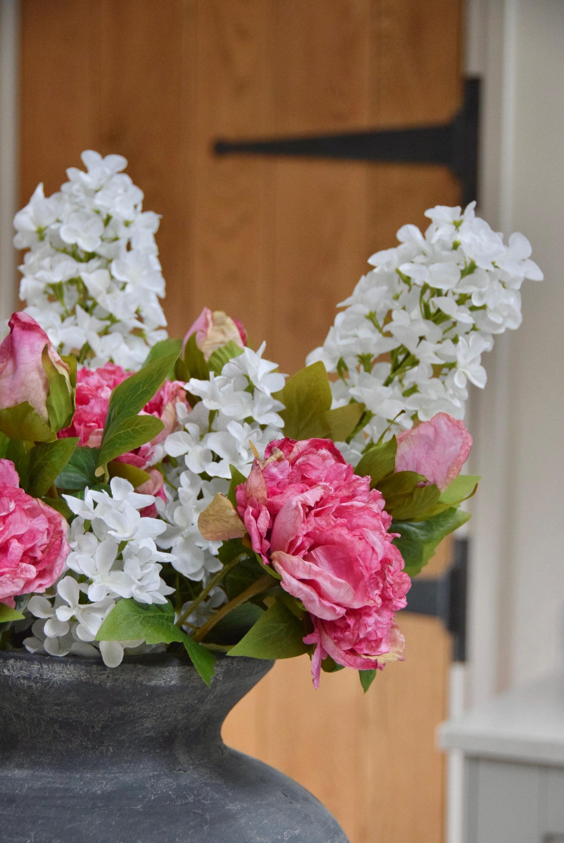 Grey antique vase placed on a kitchen countertop with an arrangement of pink peonies and white paniculata hydrangeas. Displayed in a country style kitchen for home decor.