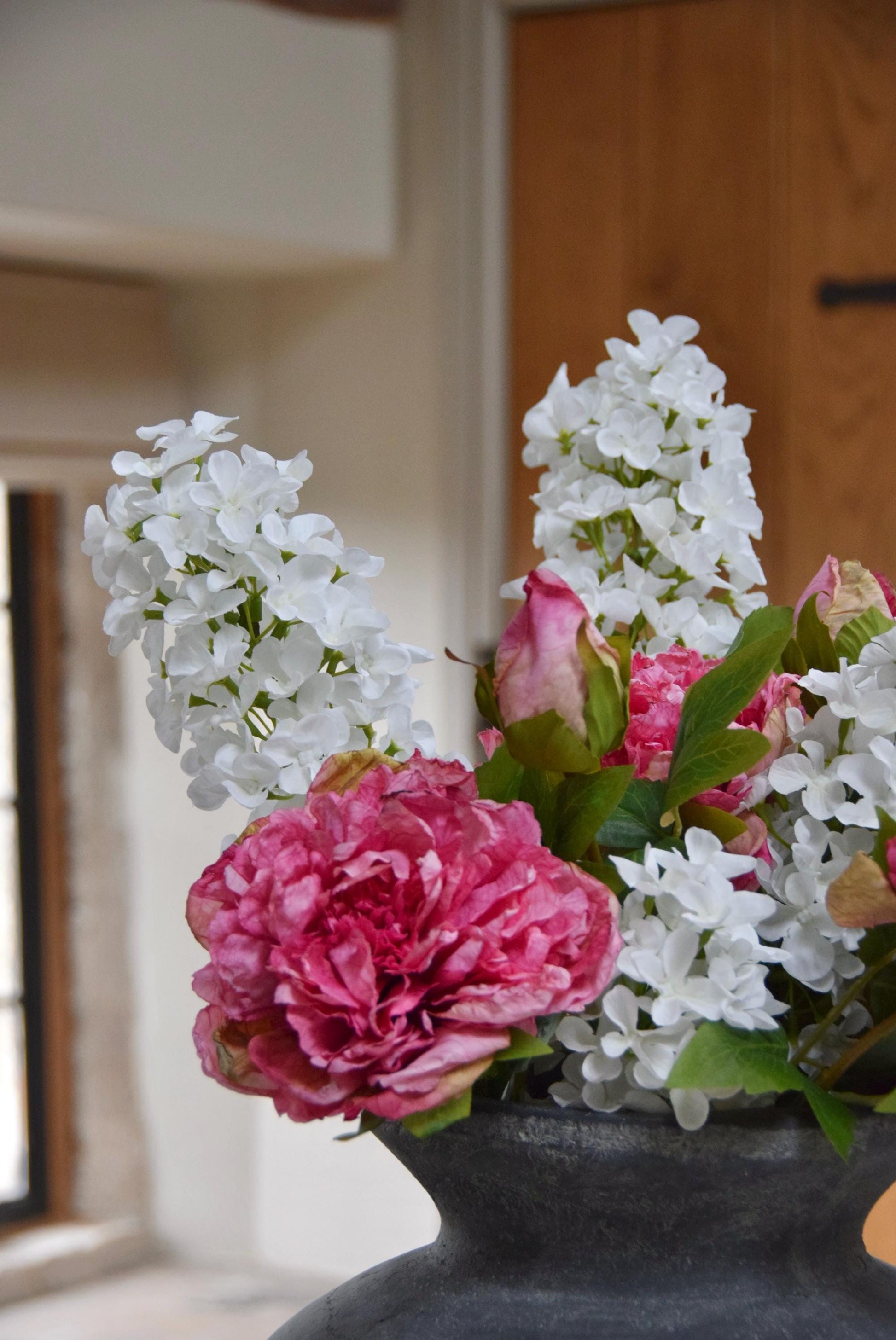 Grey antique vase placed on a kitchen countertop with an arrangement of pink peonies and white paniculata hydrangeas. Displayed in a country style kitchen for home decor.