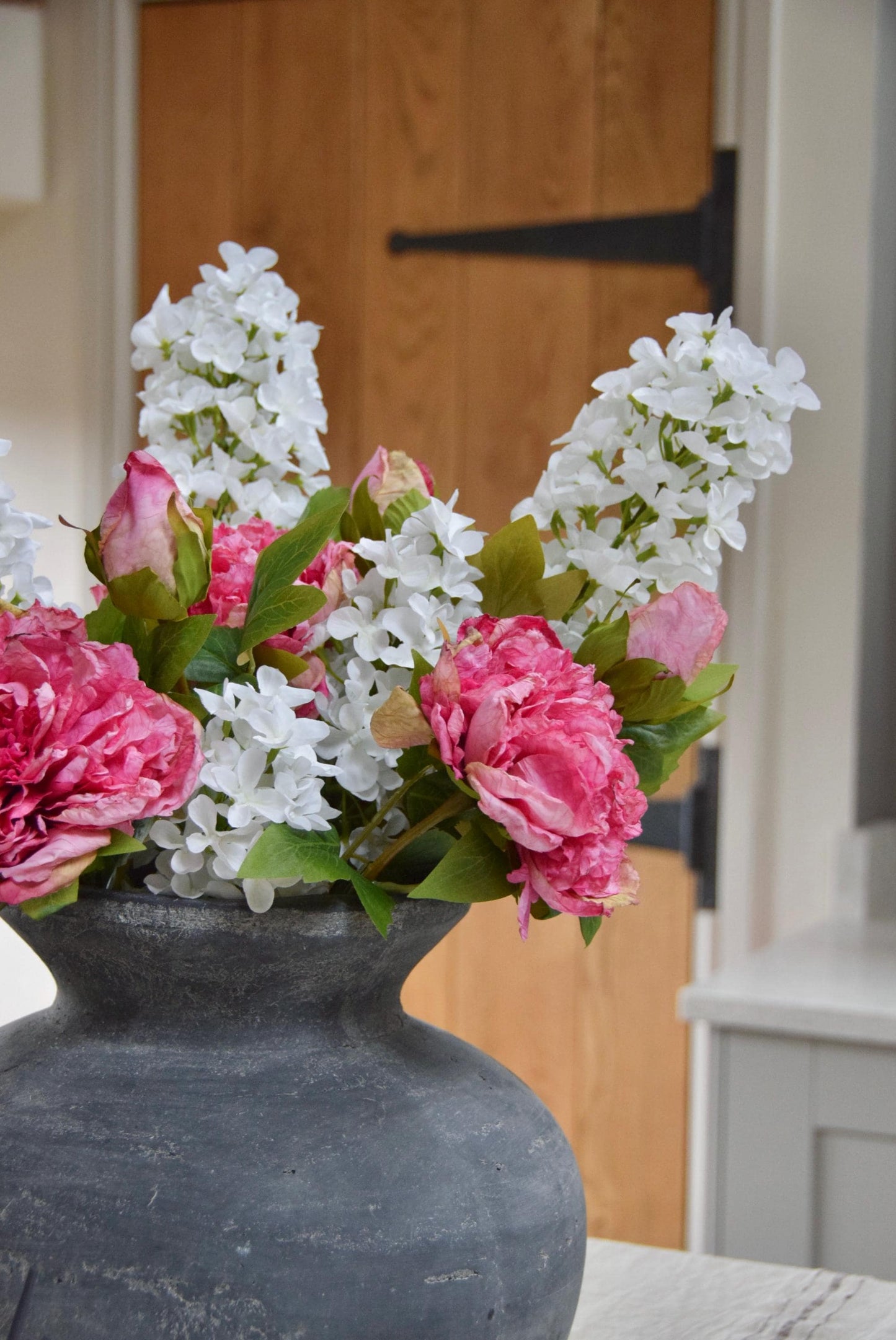 Grey antique vase placed on a kitchen countertop with an arrangement of pink peonies and white paniculata hydrangeas. Displayed in a country style kitchen for home decor.