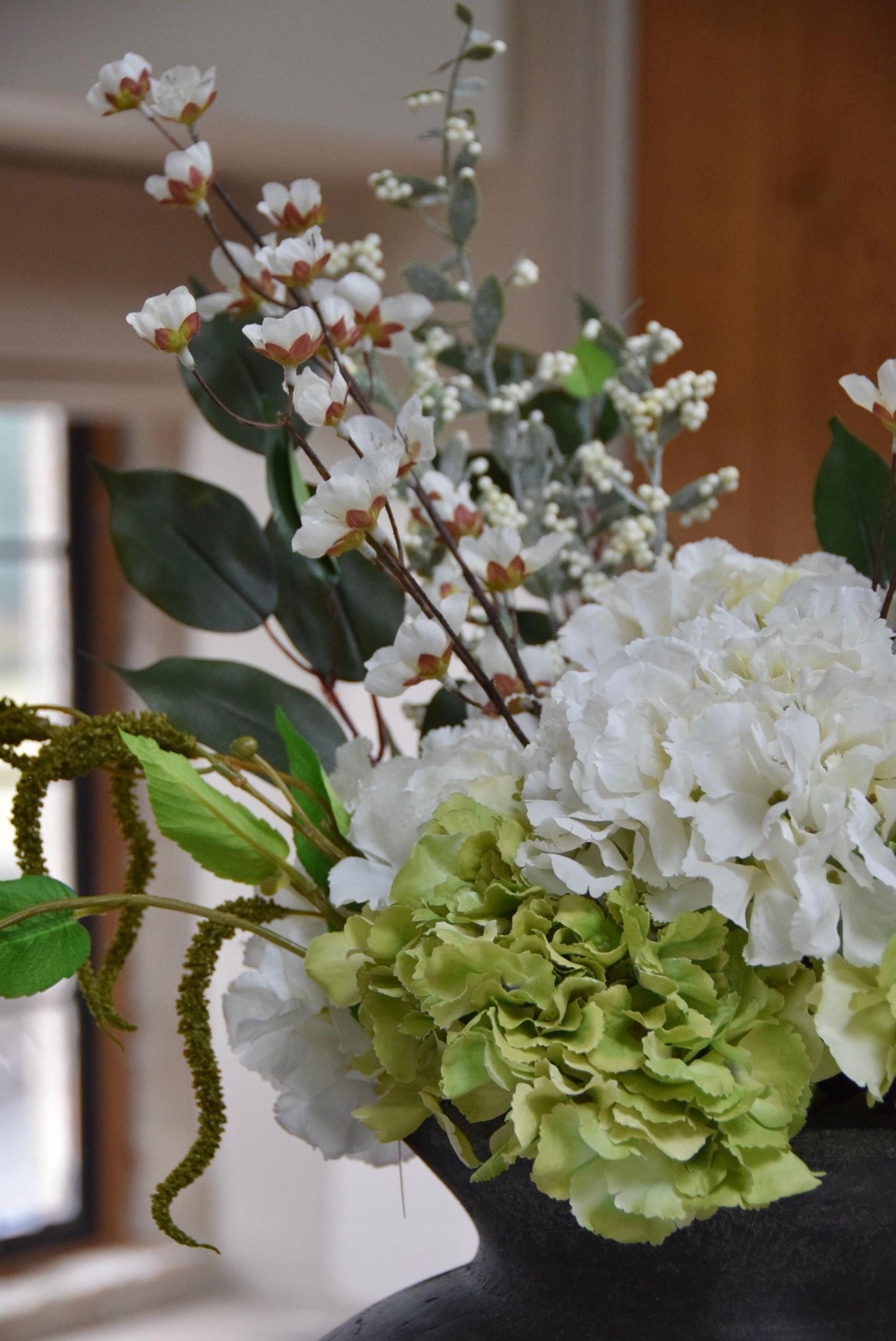 Grey vase placed on a kitchen countertop with a mixture of green and white hydrangeas with lots of mixed foliage. Displayed in a country style kitchen for home decor.