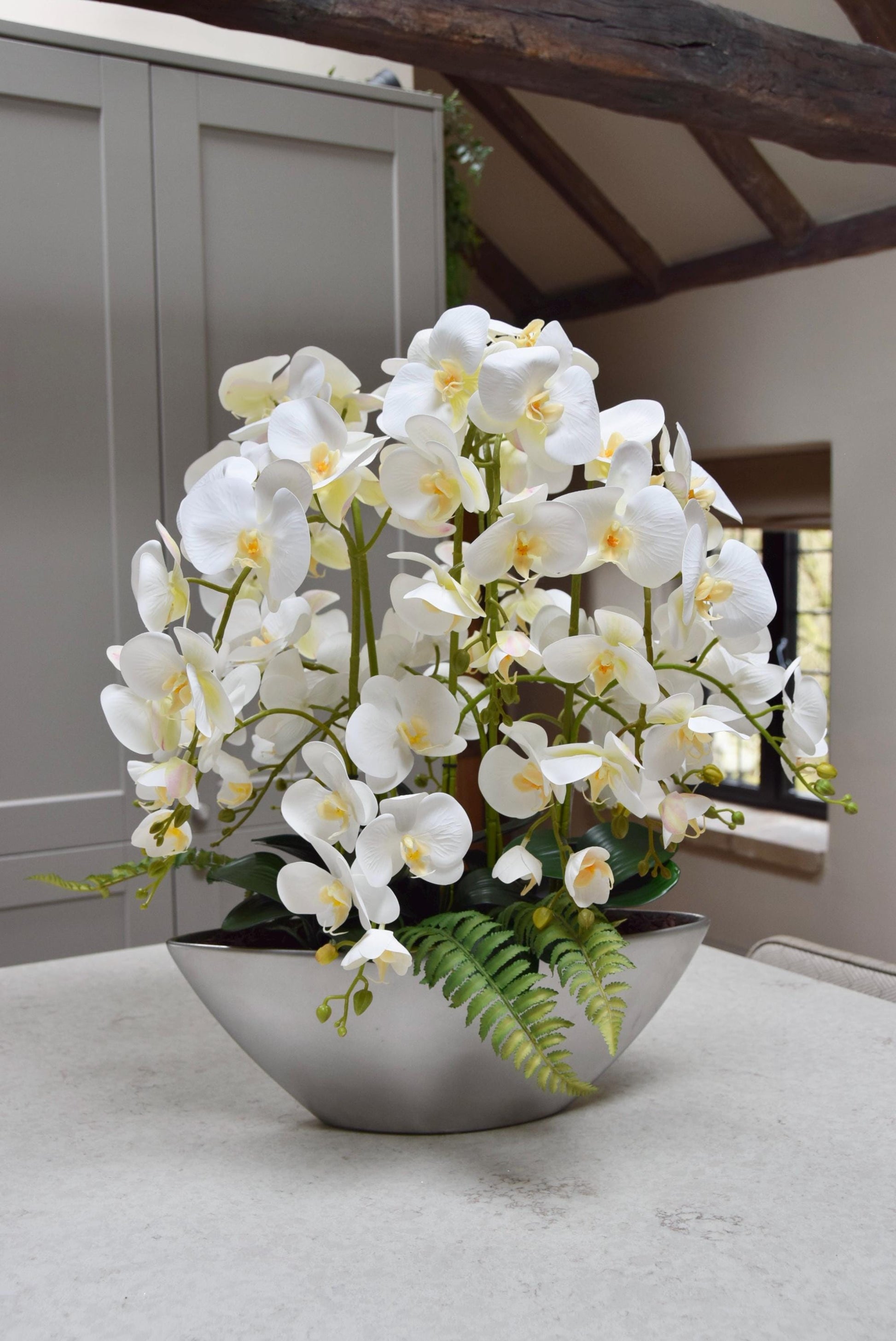Silver ark bowl placed on a kitchen counter top with an arrangement of white orchids. Arranged in a country style kitchen for home decor.
