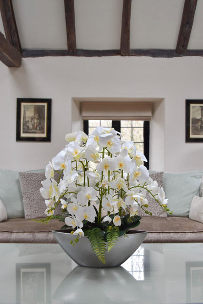 Silver ark bowl placed on a coffee table with an arrangement of white orchids. Arranged in a country style living room for home decor.