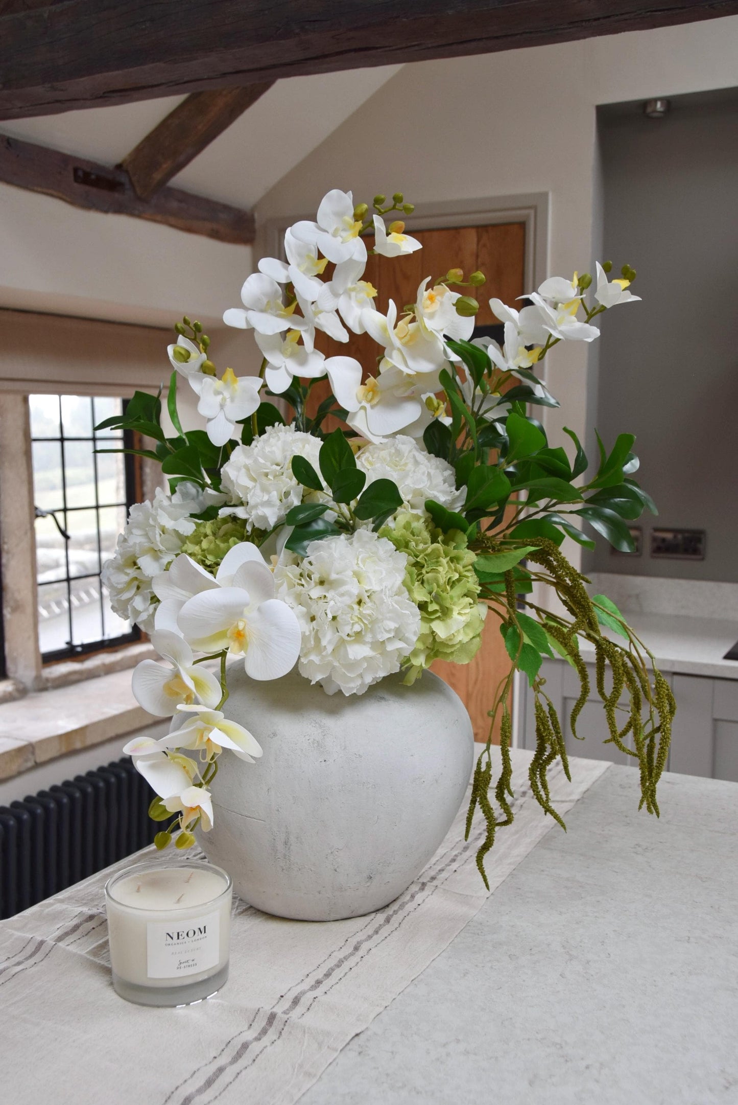 White stone vase placed on a kitchen counter top with an arrangement of white orchids, white hydrangeas, green hydrangeas and green foliage. Displayed in a country style kitchen for home decor.