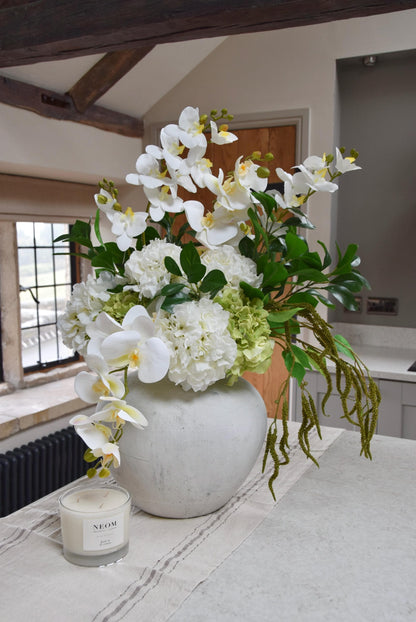 White stone vase placed on a kitchen counter top with an arrangement of white orchids, white hydrangeas, green hydrangeas and green foliage. Displayed in a country style kitchen for home decor.