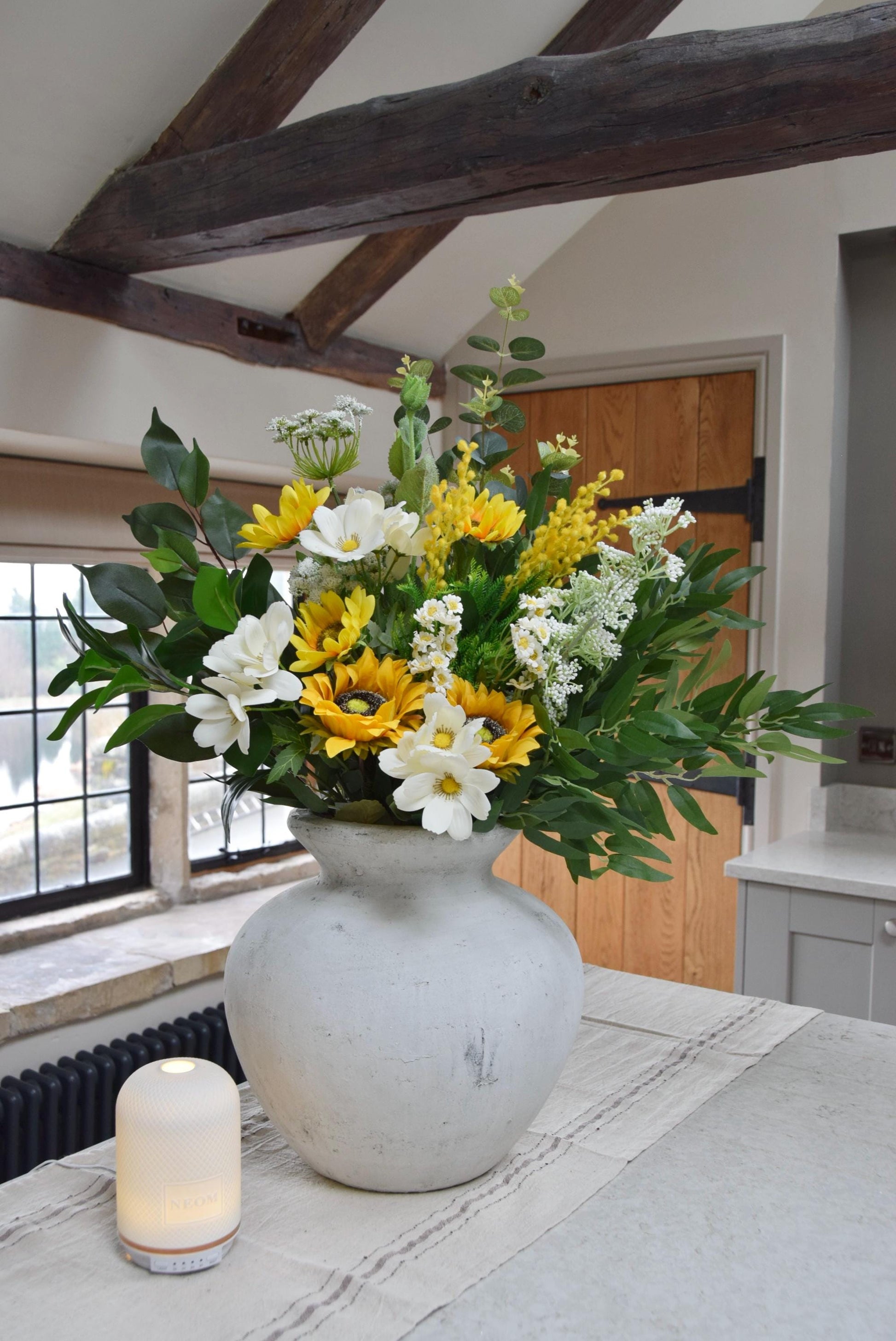 White stone vase placed on a kitchen countertop with a mixture of yellow sunflowers, white wildflowers and foliage. Displayed in a country style kitchen for home decor.