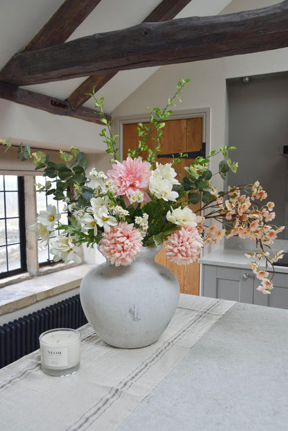 White stone vase placed on a kitchen countertop with a mixture of pink mum flowers, pink dahlias, white wildflowers and green foliage. Displayed in a country style kitchen for home decor.
