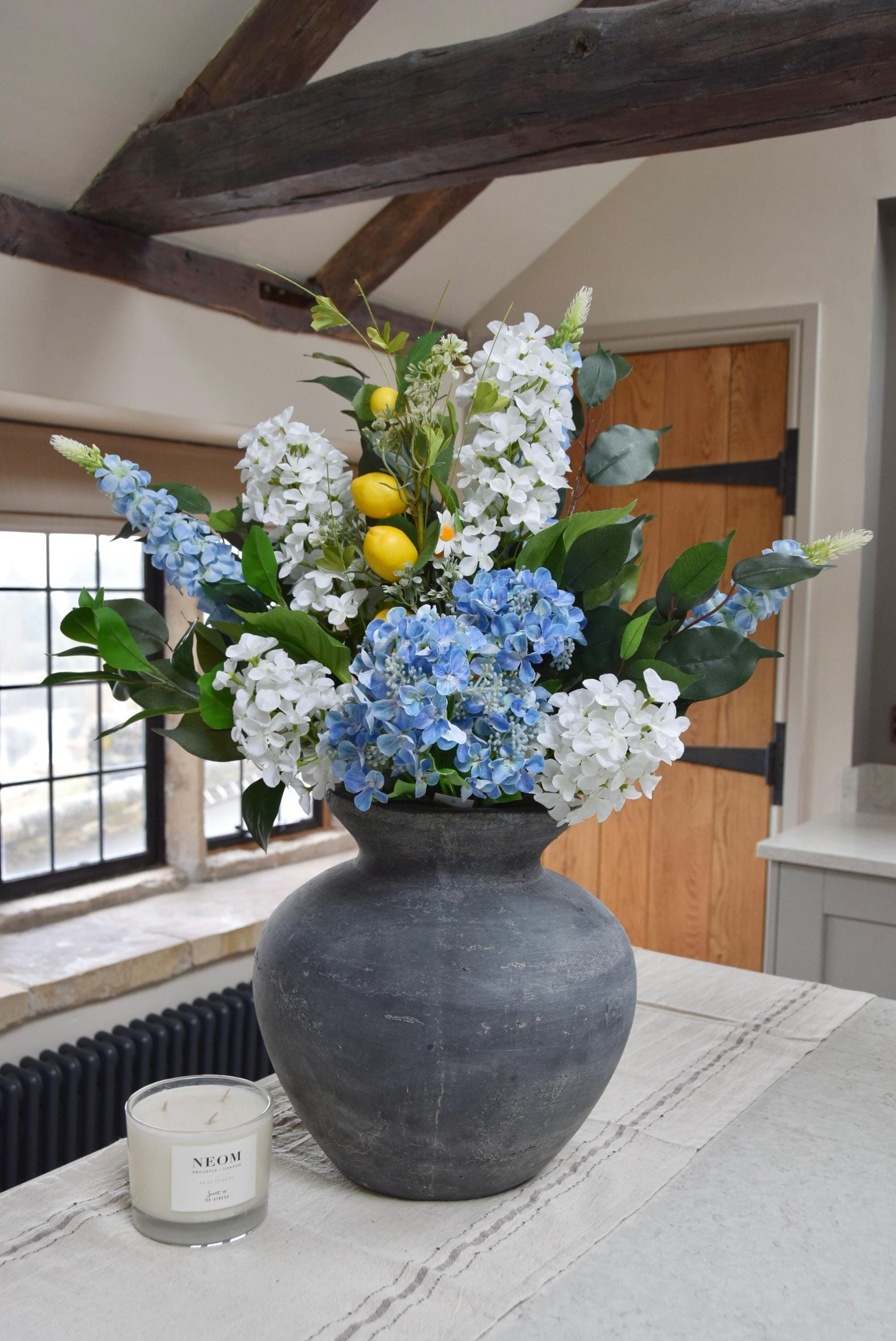 Grey antique vase placed on a kitchen counter top with blue and white hydrangeas and lemon branches. Displayed in a country style kitchen for home decor.