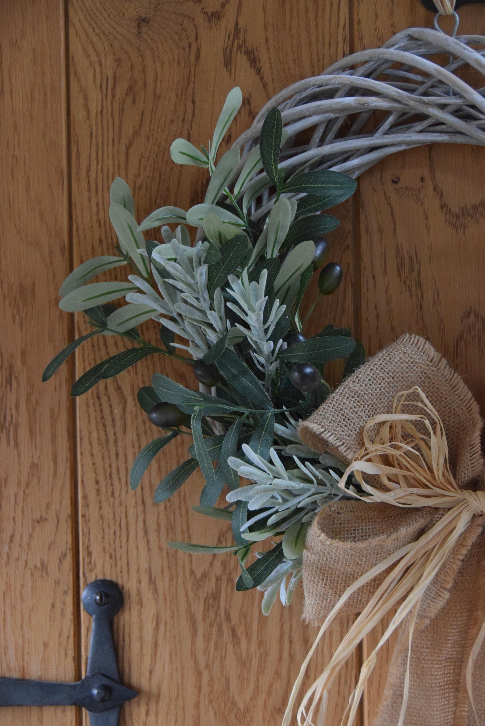 Olive branch wreath created on a willow wreath hanging on a country style door. Mixed with olive branches and tarragon herbs, finished with raffia and natural ribbon.