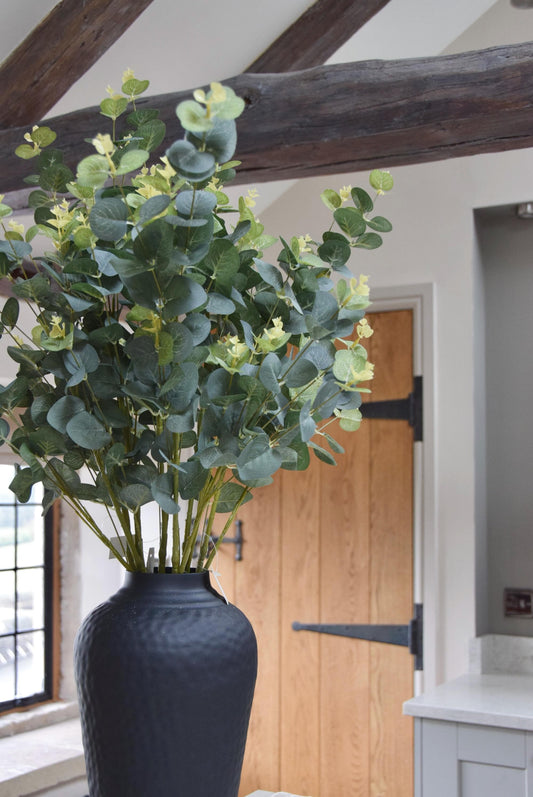 Tall black vase placed on a kitchen countertop with a bunch of green artificial eucalyptus. Displayed in a country style kitchen for home decor.