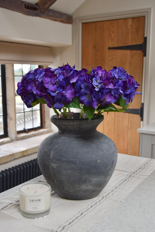 Grey antique vase placed on a kitchen countertop with a bunch of artificial purple hydrangeas. Displayed in a country style kitchen for home decor.