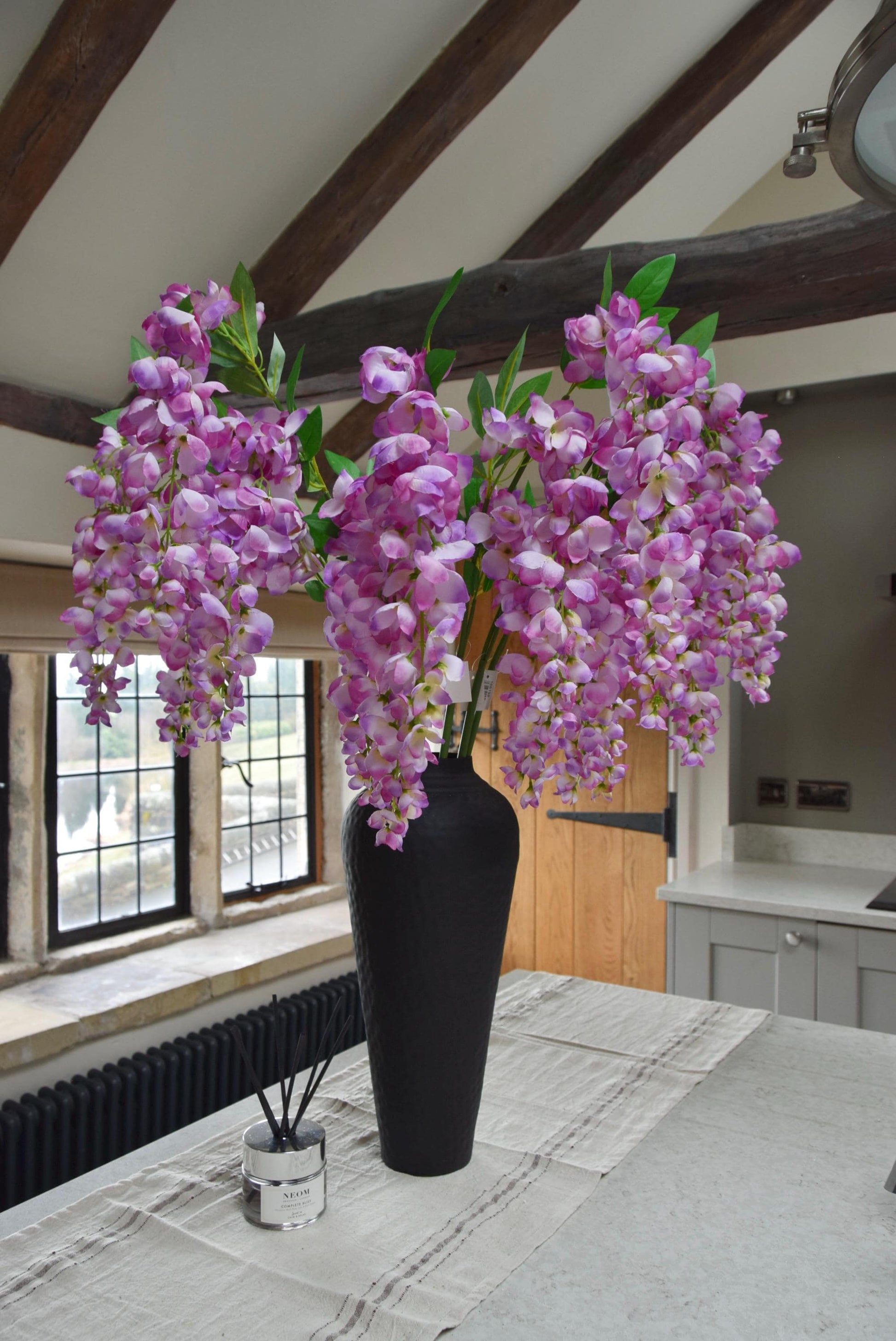 Tall black vase set on a kitchen countertop with a bunch of purple artificial wisteria stems. Displayed in a country style kitchen for home decor.