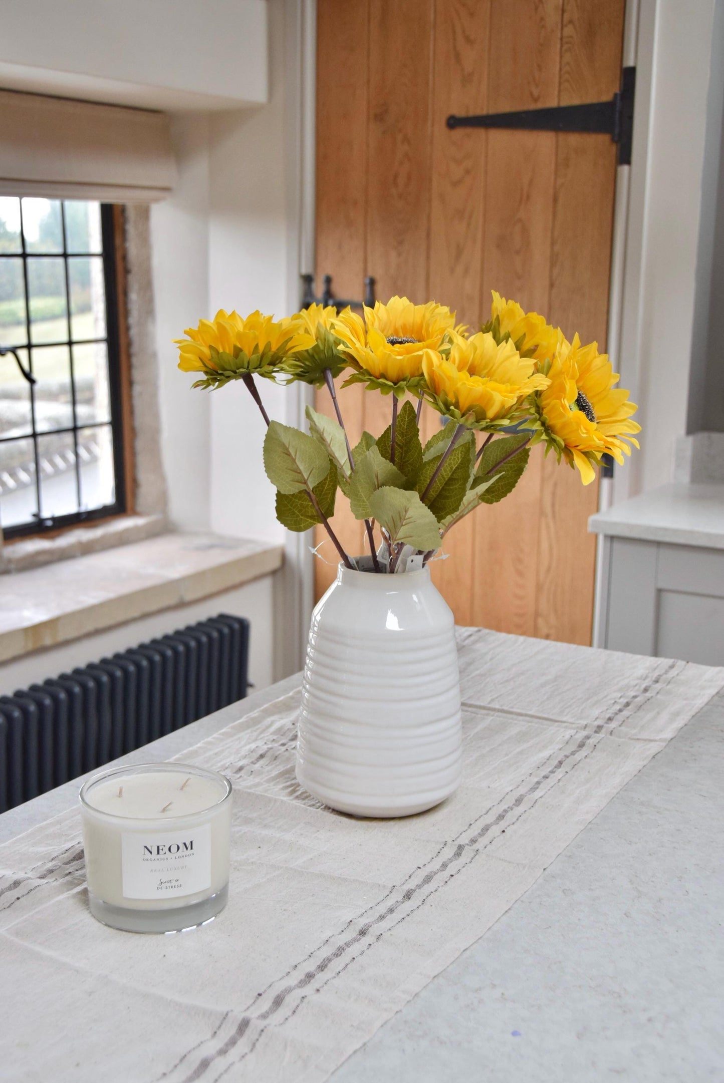 Artificial yellow sunflowers set in a cream ceramic vase placed on a kitchen countertop. Displayed in a country style kitchen for home decor.