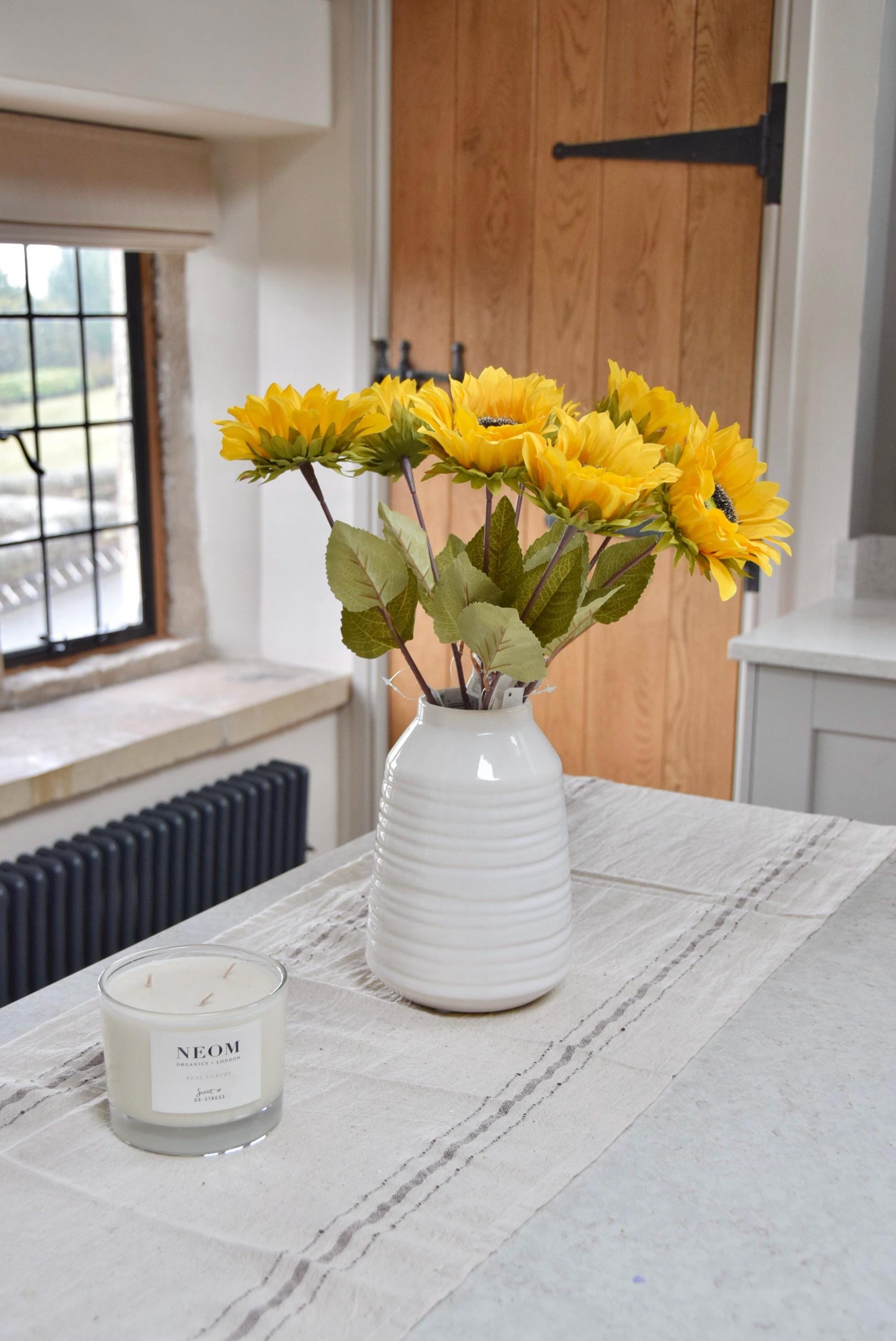 Artificial yellow sunflowers set in a cream ceramic vase placed on a kitchen countertop. Displayed in a country style kitchen for home decor.