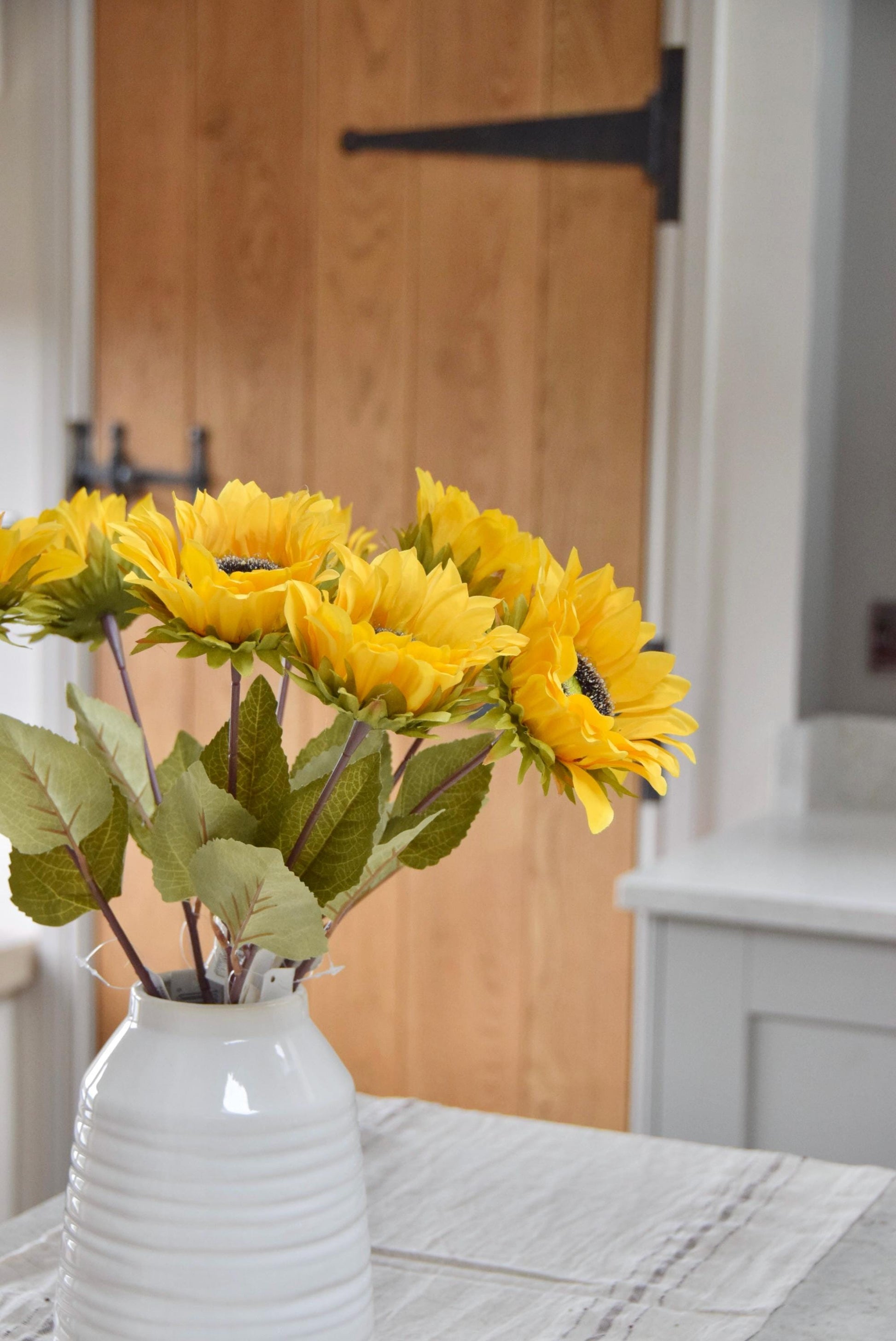 Artificial yellow sunflowers set in a cream ceramic vase placed on a kitchen countertop. Displayed in a country style kitchen for home decor.