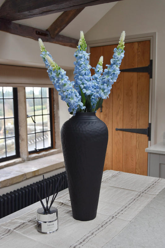 Tall black vase placed on a kitchen countertop with a bunch of blue delphiniums. Displayed in a country style kitchen for home decor.