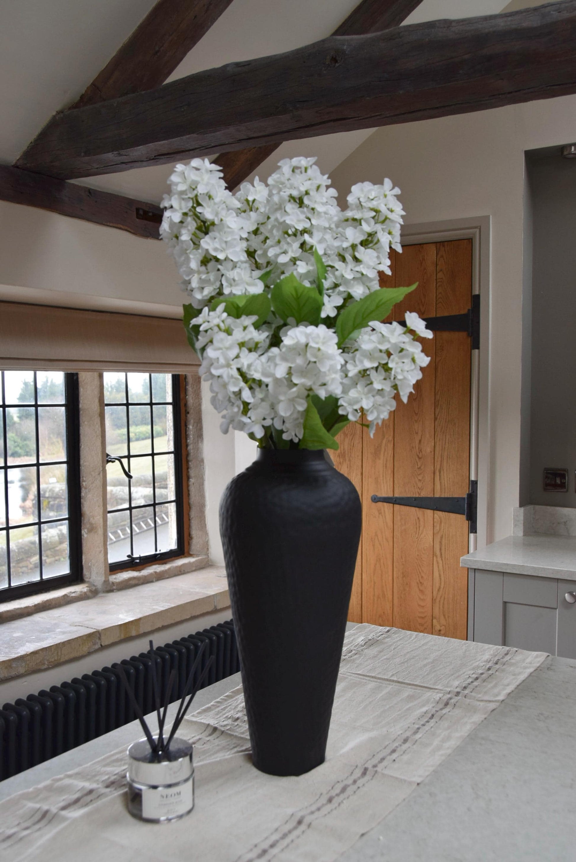 Tall black vase placed on a kitchen countertop with a bunch of paniculata hydrangeas. Displayed in a country style kitchen for home decor.