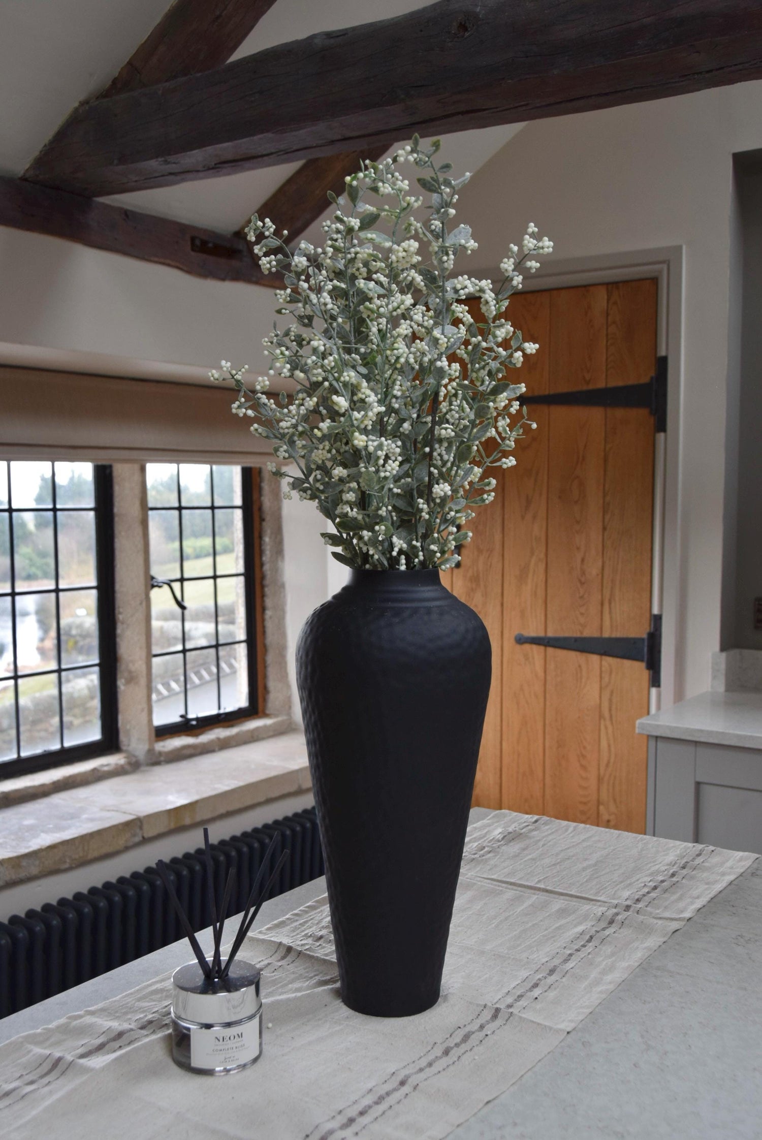 Tall black vase placed on a kitchen countertop with a bunch of artificial berry leaf stems. Displayed in a country style kitchen for home decor.
