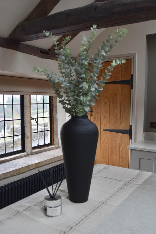 Tall black vase placed on a kitchen countertop with a bunch of dusted artificial eucalyptus stems. Displayed in a country style kitchen for home decor.