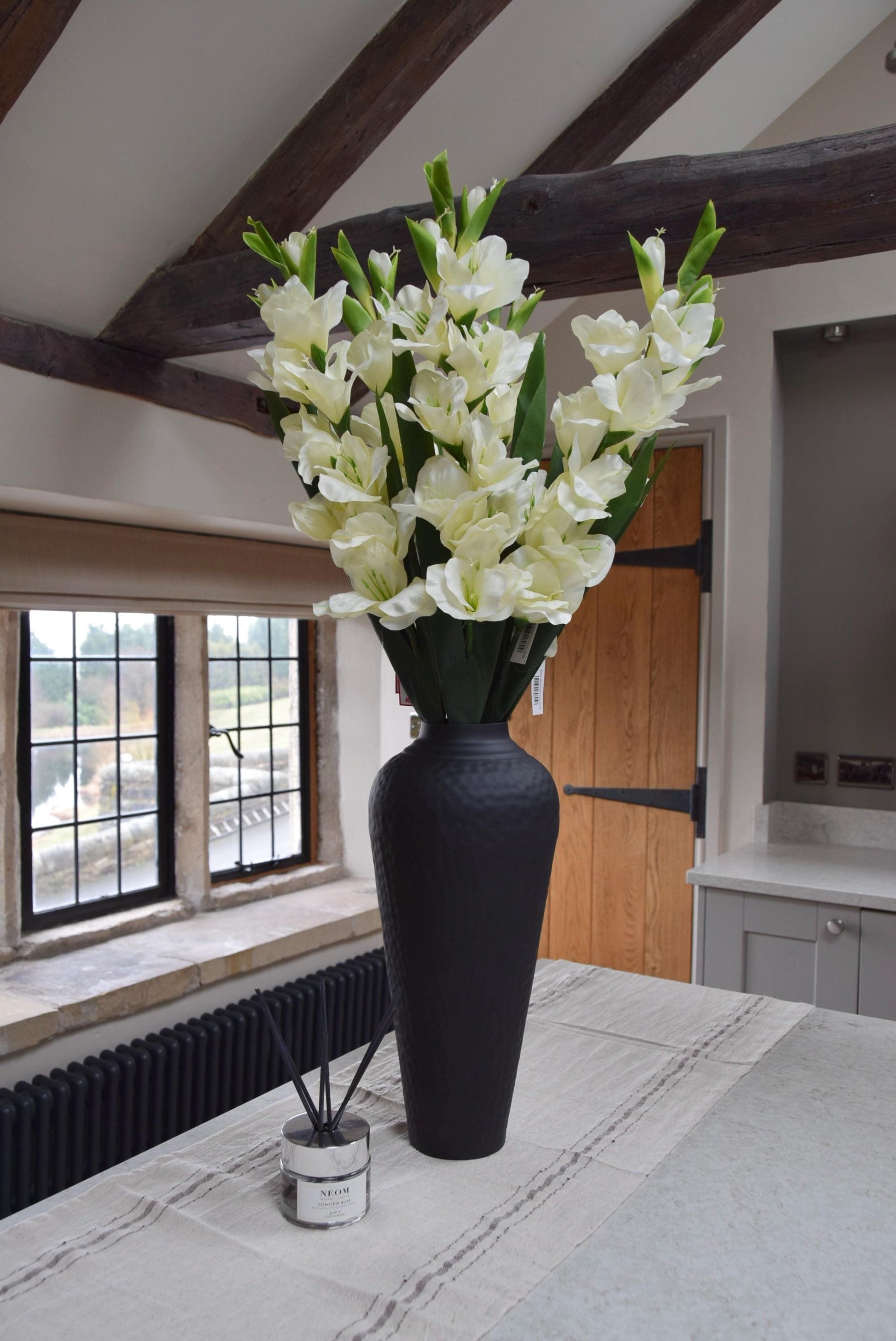 Tall black vase placed on a kitchen countertop with a bunch of gladioli stems. Displayed in a country style kitchen for home decor.