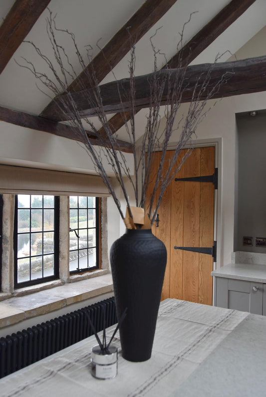 Tall black vase placed on a kitchen countertop with a bunch of faux brown branches. Displayed in a country style kitchen for home decor.