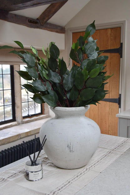 Stone antique vase placed on a kitchen countertop with a bunch of artificial bay leaf sprays. Displayed in a country style kitchen for home decor.