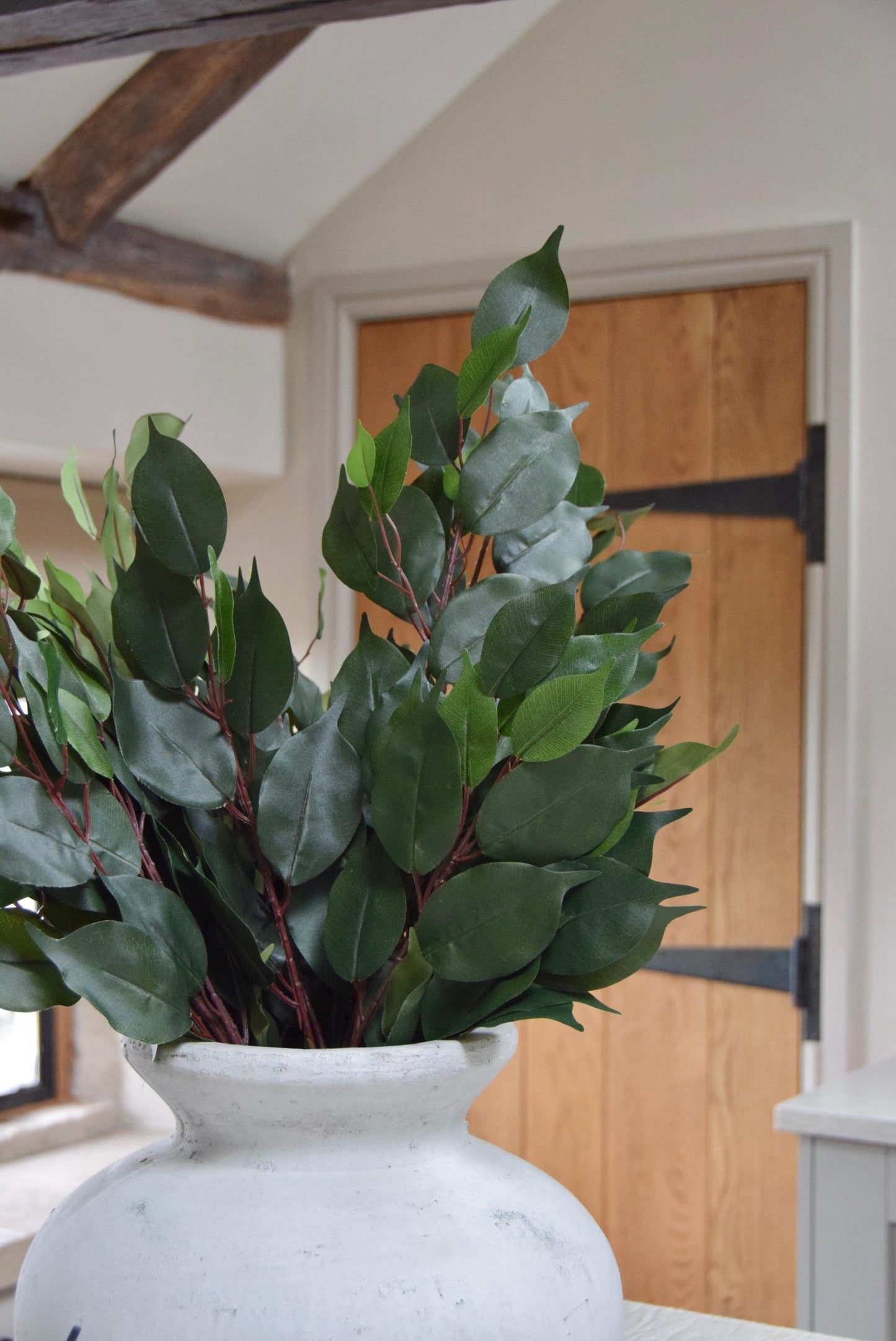 Stone antique vase placed on a kitchen countertop with a bunch of artificial bay leaf sprays. Displayed in a country style kitchen for home decor.