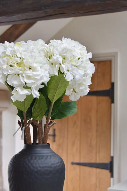 Tall black vase placed on a kitchen countertop with white hydrangeas. Displayed in a country style kitchen for home decor.