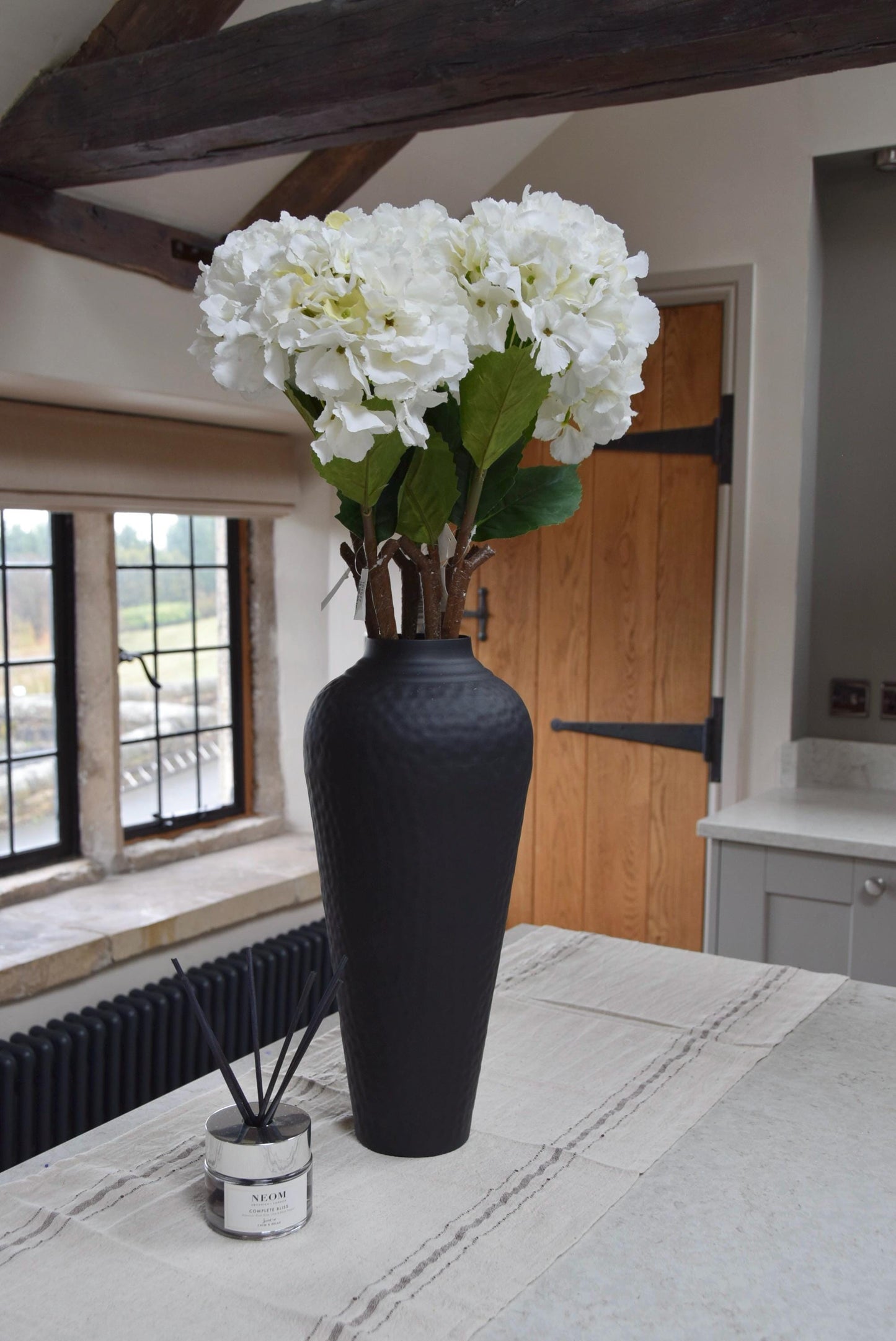 Tall black vase placed on a kitchen countertop with white hydrangeas. Displayed in a country style kitchen for home decor.