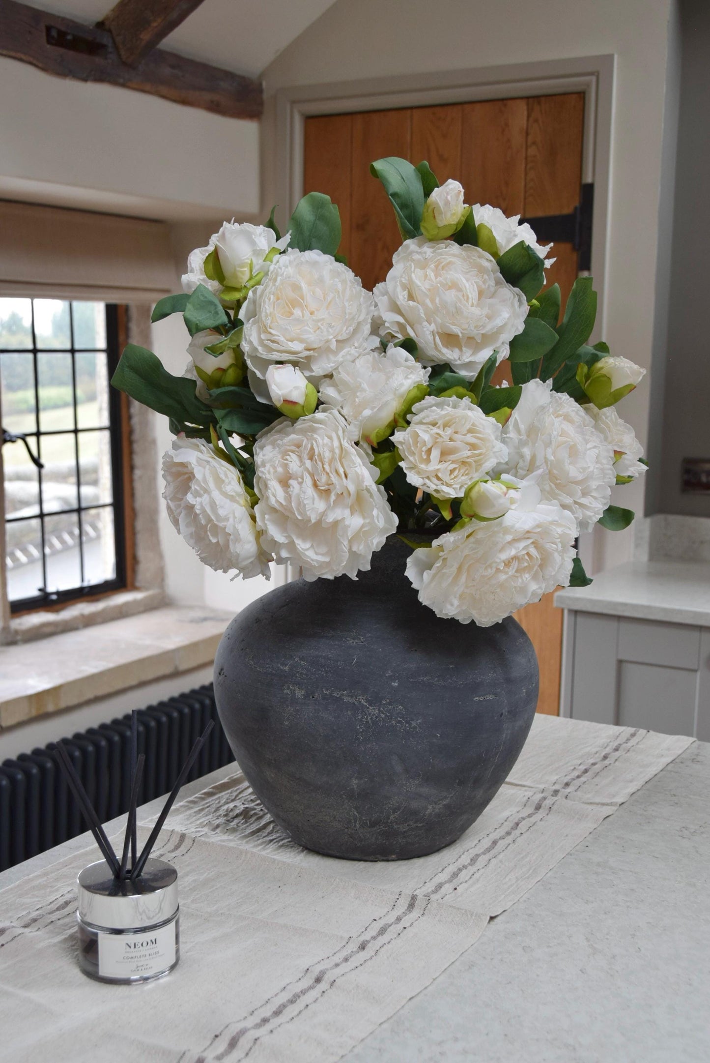 Grey antique vase placed on a kitchen countertop with a bunch of artificial cream peonies. Displayed in a country style kitchen for home decor.