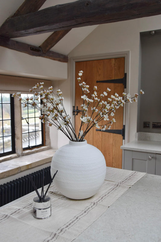 Round white vase placed on a kitchen countertop with a bunch of artificial cherry blossom branches. Displayed in a country style kitchen for home decor.