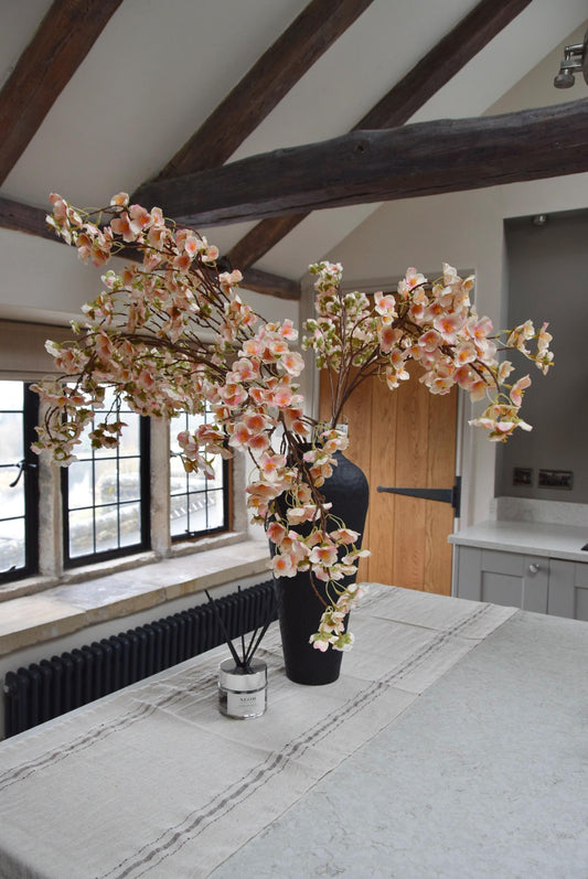 Tall black vase placed on a kitchen countertop with a bunch of hanging pink plum blossom stems. Displayed in a country style kitchen for home decor.
