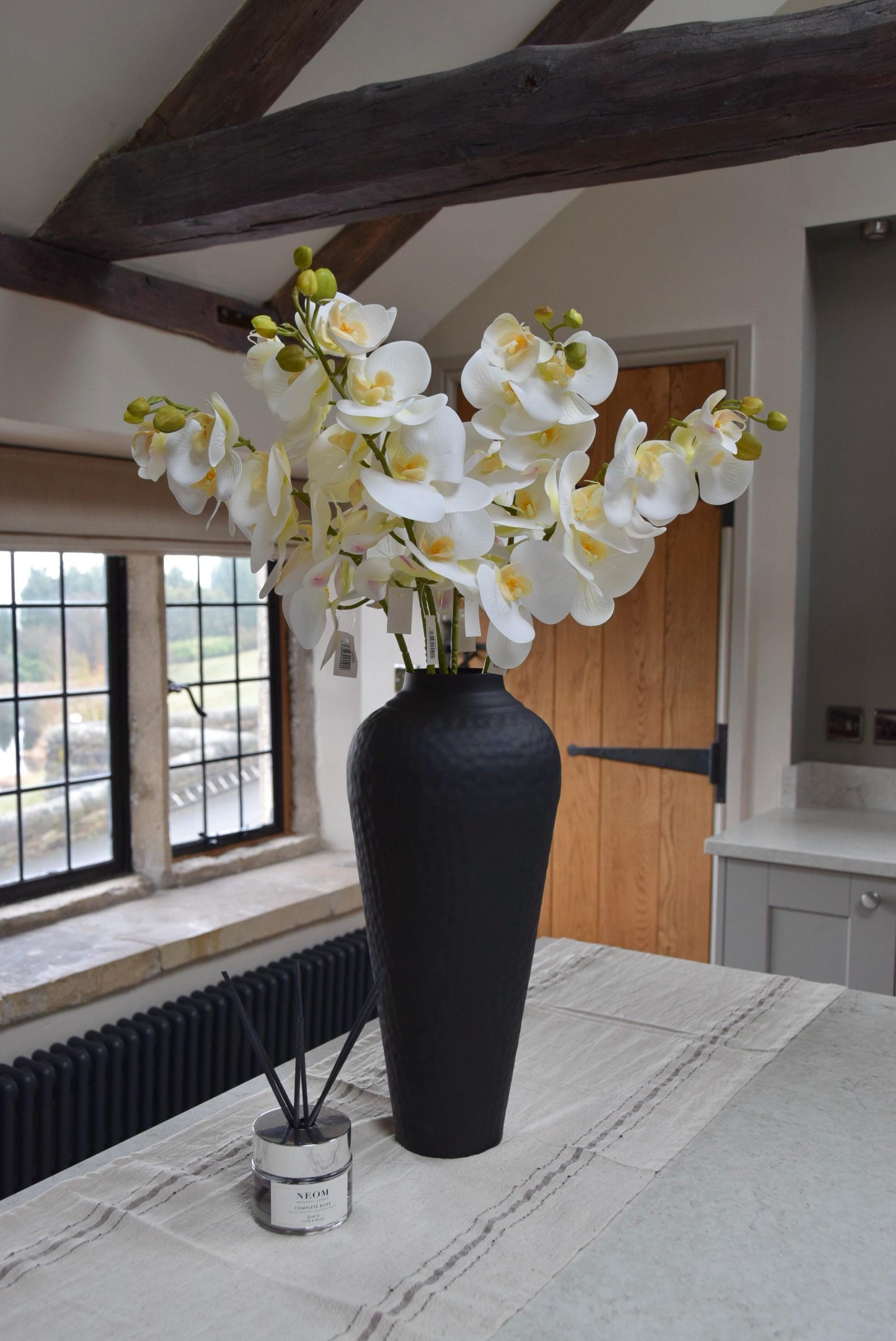 Tall black vase placed on a kitchen countertop with a bunch of white orchid stems. Displayed in a country style kitchen for home decor.