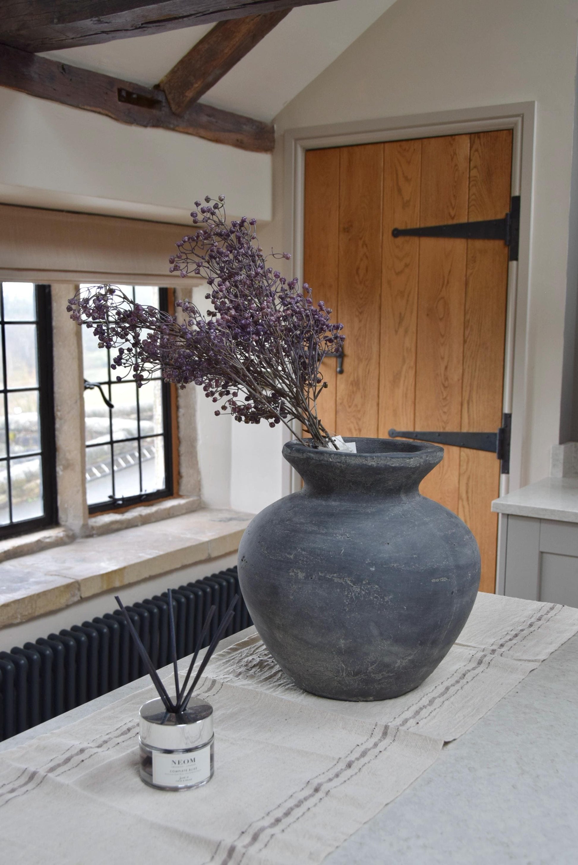 Grey antique vase with a bunch of purple dried look linum sprays placed on a kitchen countertop. Displayed in a country style kitchen for home decor.