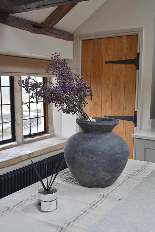 Grey antique vase with a bunch of purple dried look linum sprays placed on a kitchen countertop. Displayed in a country style kitchen for home decor.