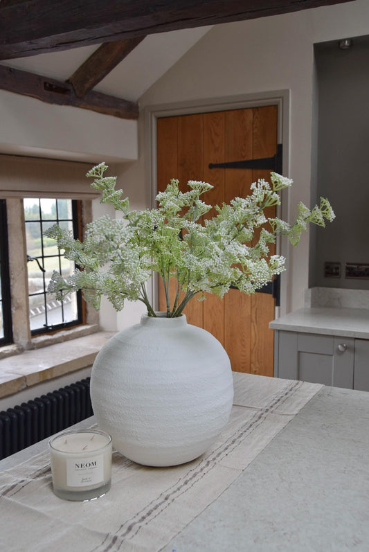 White round vase placed on a kitchen countertop with a bunch of white mini bud stems displayed in a country style kitchen for home decor.