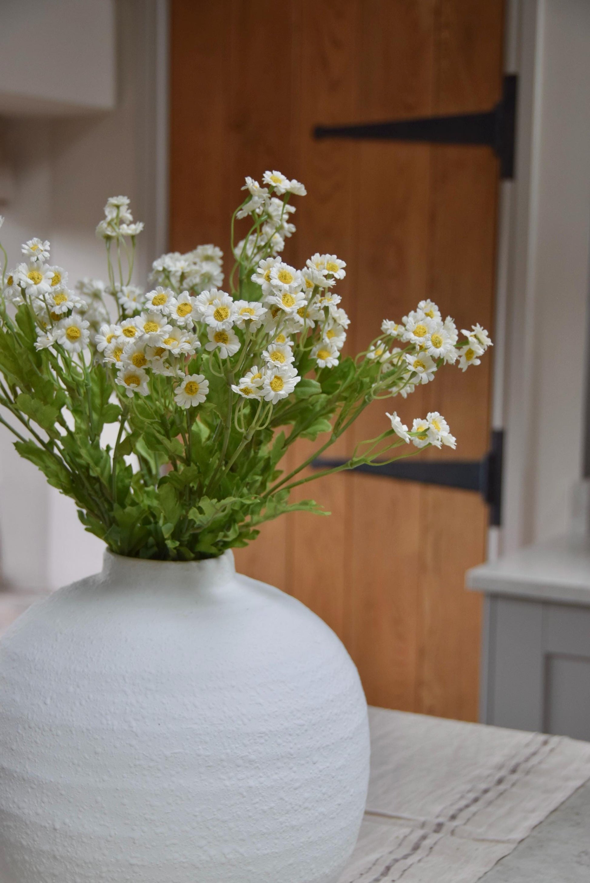 White round vase placed on a kitchen countertop with a bunch of daisies displayed in a country style kitchen for home decor.