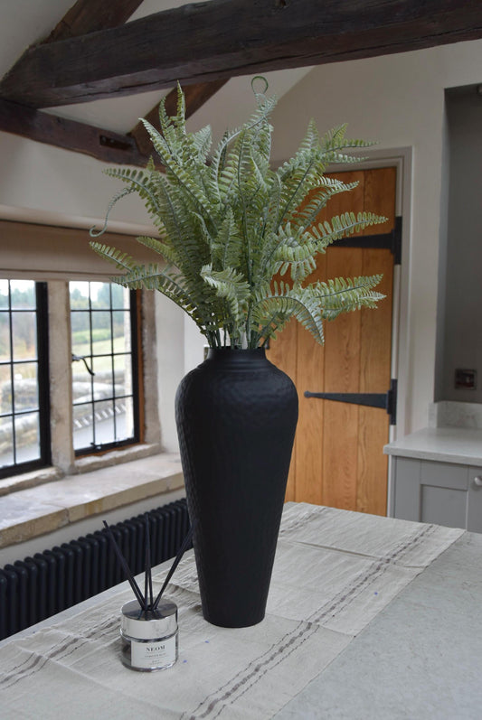 Tall black vase with long dusted fern stems placed on a kitchen countertop in a country style house.
