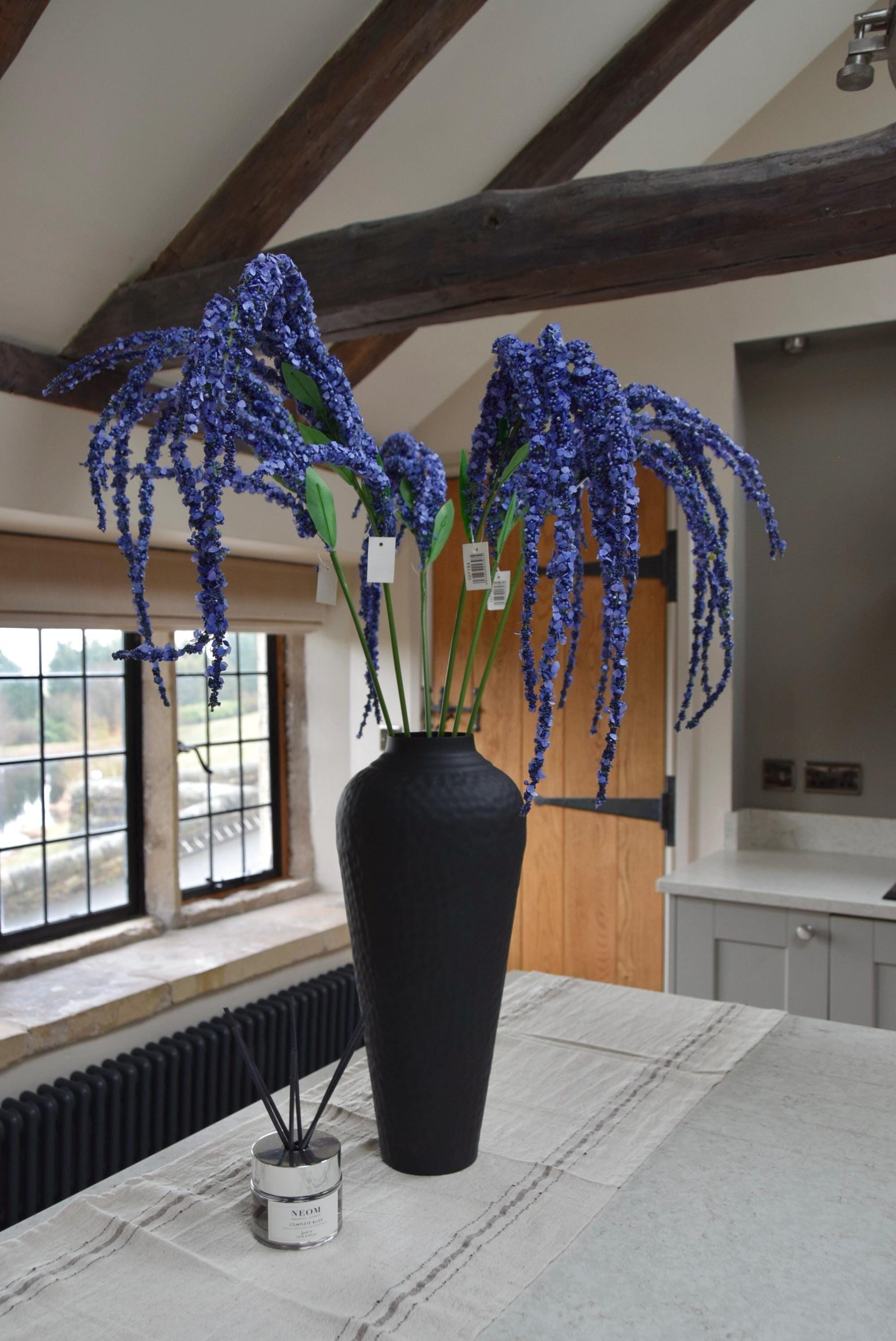 Tall black vase on a kitchen countertop with long purple amaranthus trailing stems. Set in a country style kitchen for home decor.